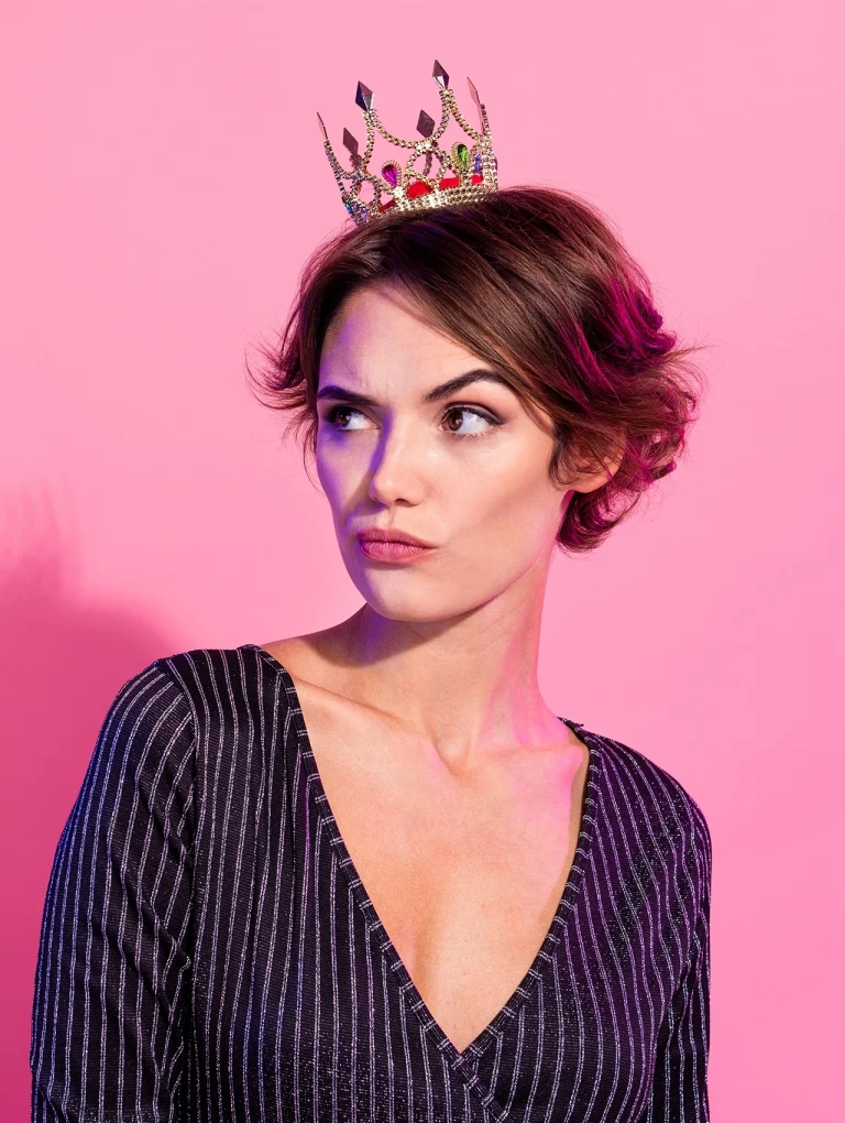 Mujer narcisista con corona sobre fondo rosa mirando de lado.
