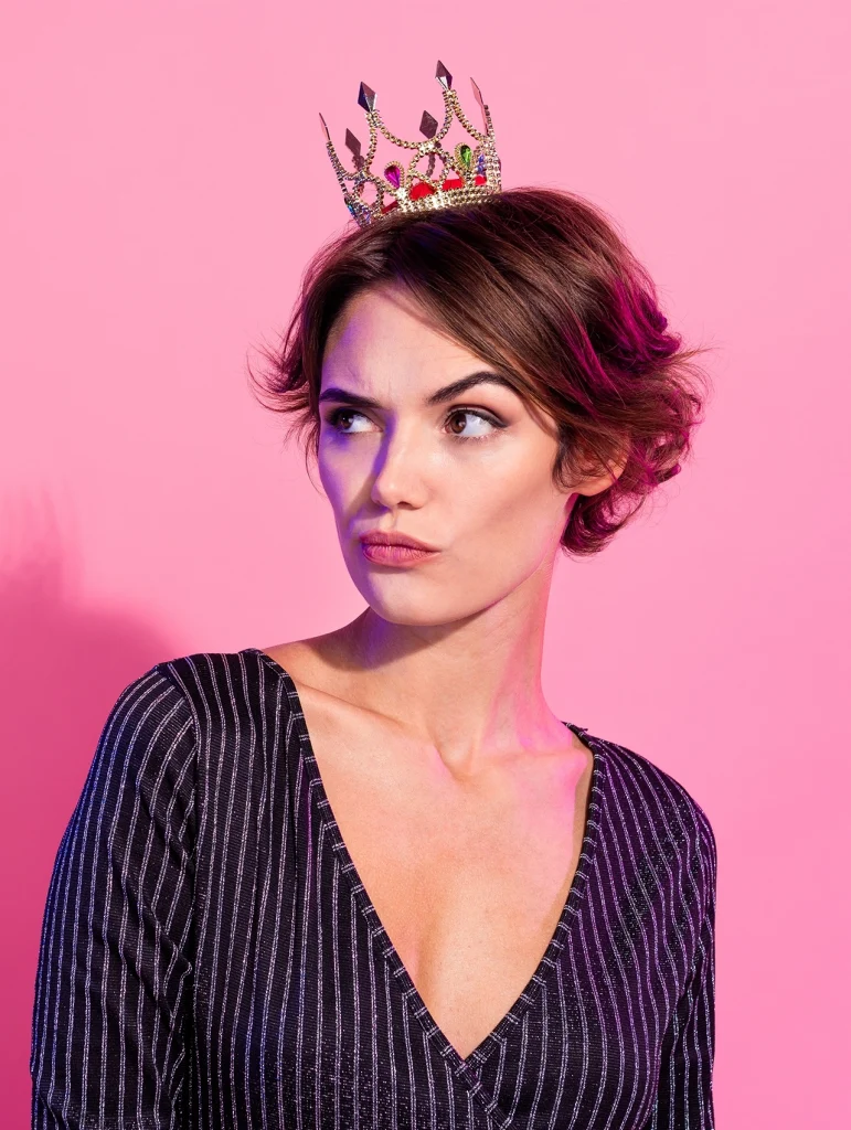 Mujer narcisista con corona sobre fondo rosa mirando de lado.