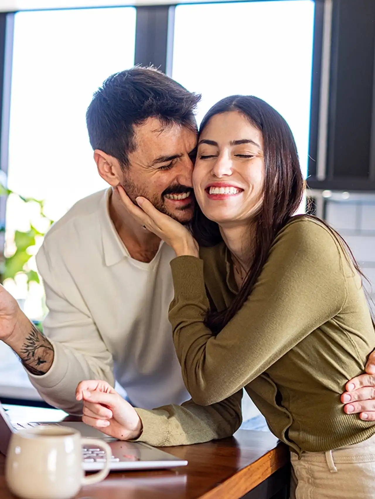 Pareja feliz abrazándose en la cocina mientras usan una laptop, mostrando el apego en las relaciones de pareja.