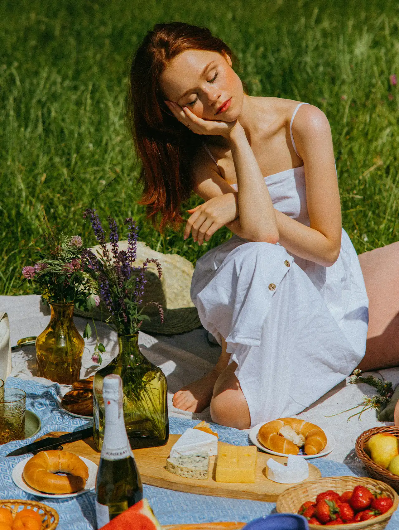 Mujer con vestido blanco disfrutando de un picnic romántico con quesos, vino espumoso y flores en el campo