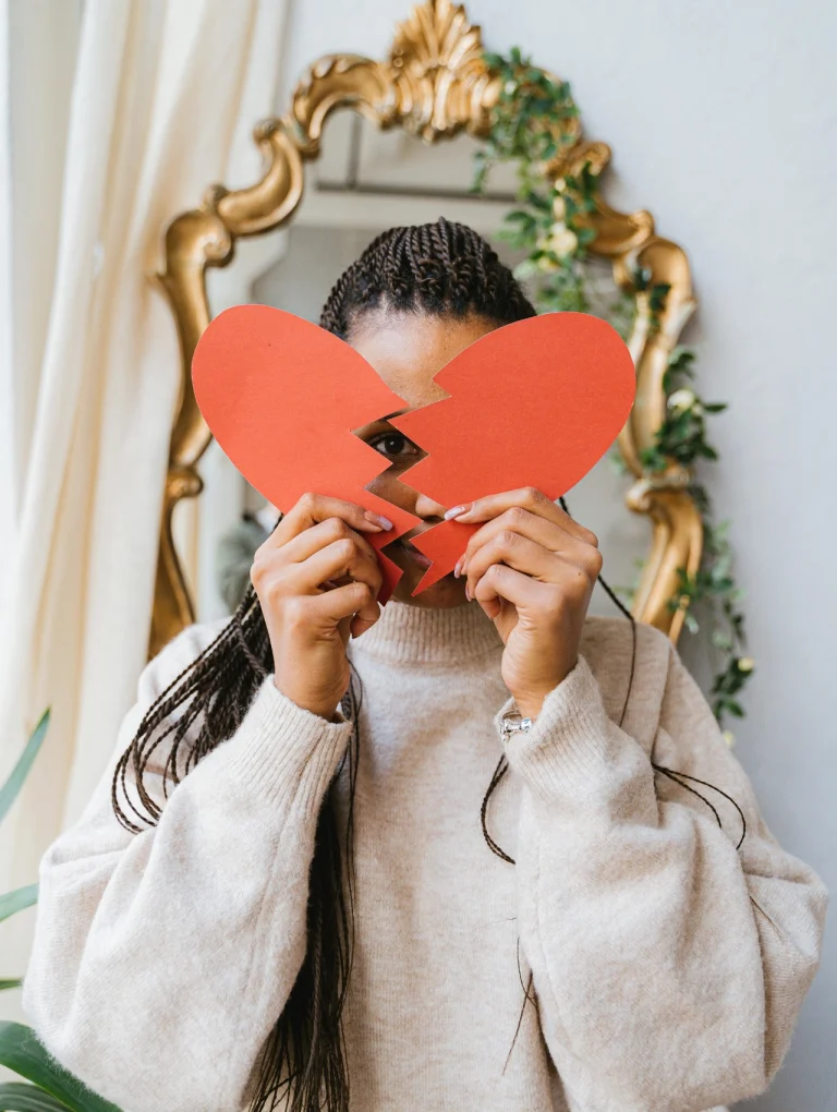 Mujer cubriendo su rostro con un corazón roto de papel, simbolizando el duelo y la resiliencia, cómo superar una ruptura amorosa.