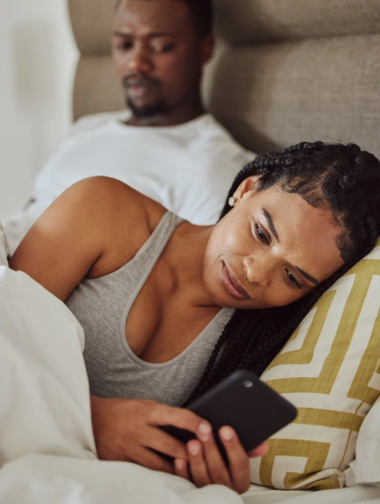Mujer acostada revisando su celular mientras su pareja la observa con desconfianza – tipos de infidelidad.