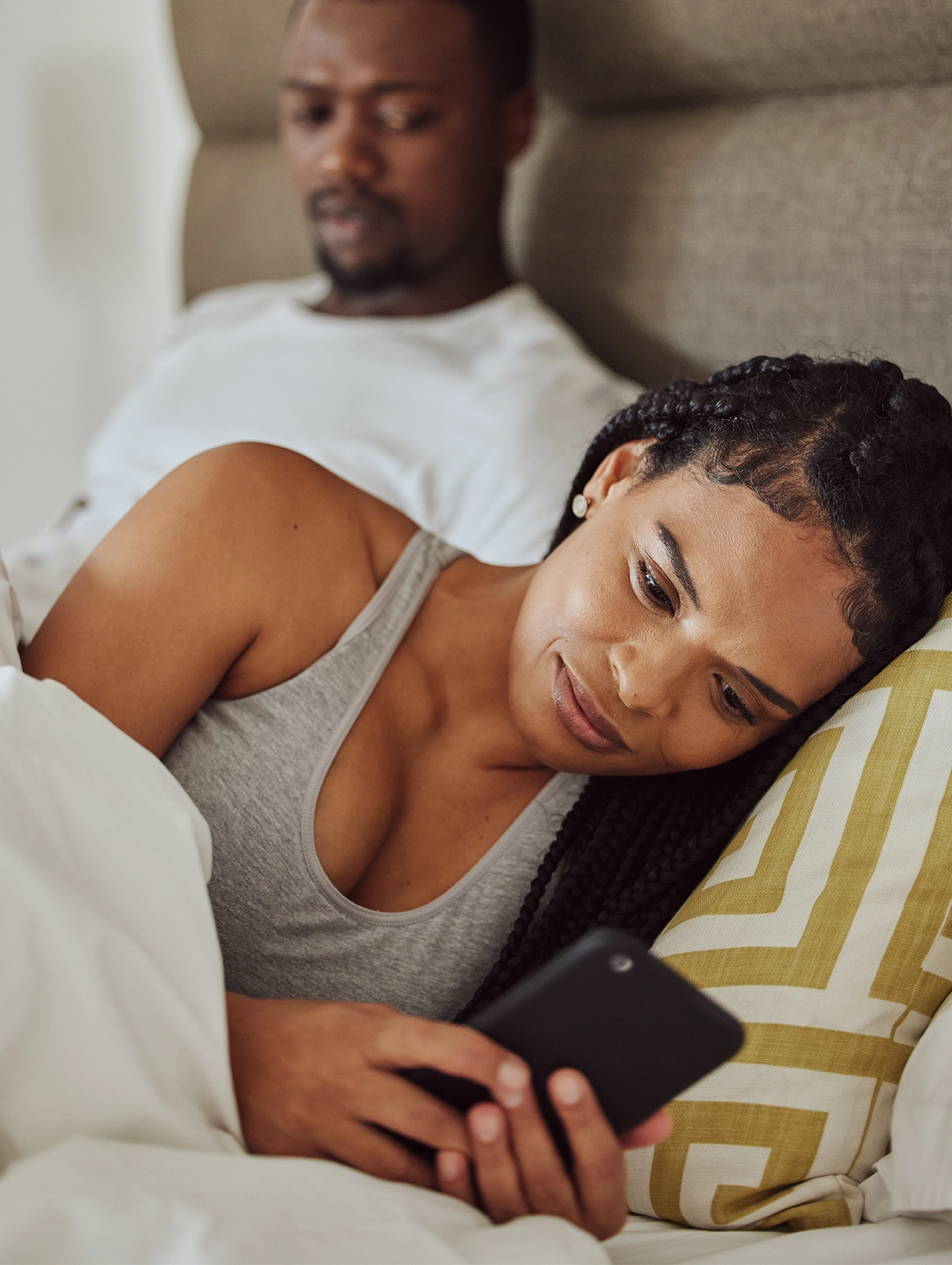 Mujer acostada revisando su celular mientras su pareja la observa con desconfianza – tipos de infidelidad.