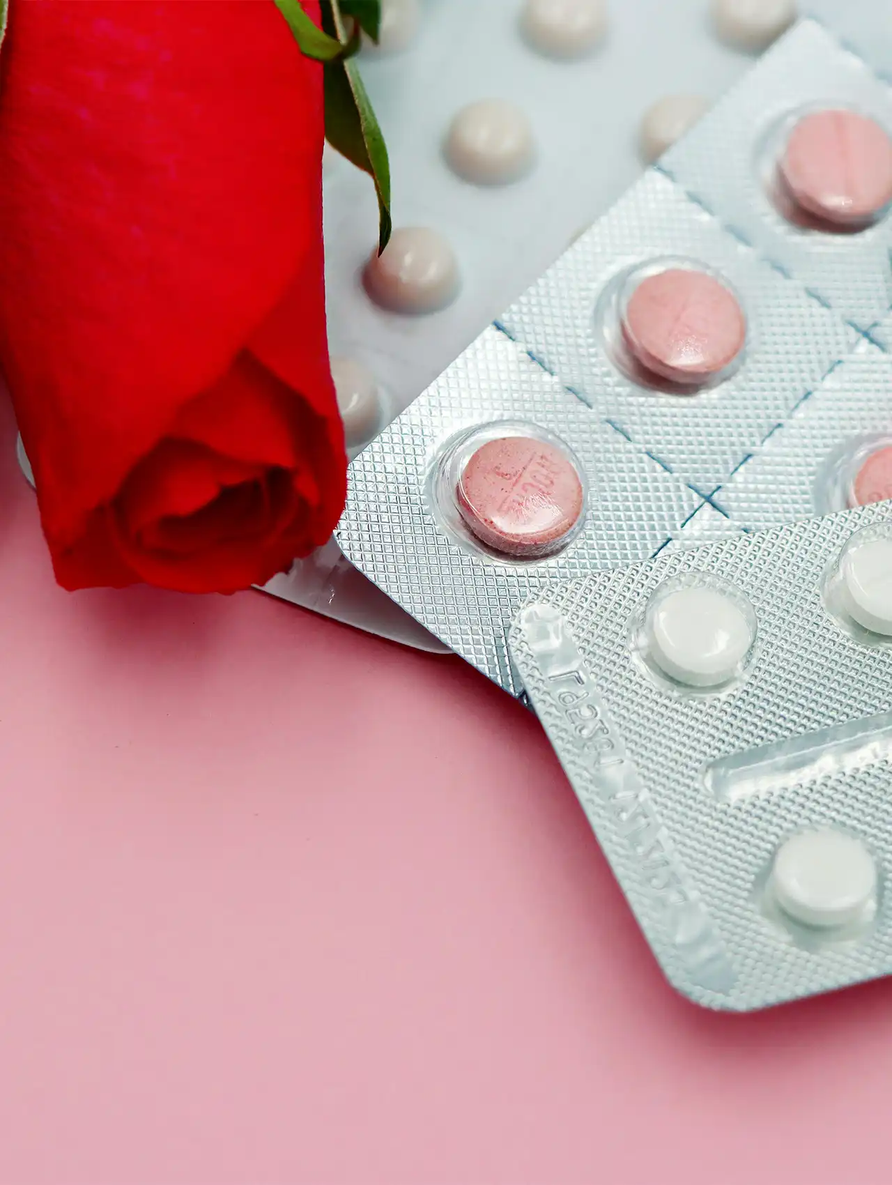 Blíster de pastillas rosadas y blancas junto a una rosa roja sobre fondo rosa, ilustración del tema las pastillas anticonceptivas son seguras.