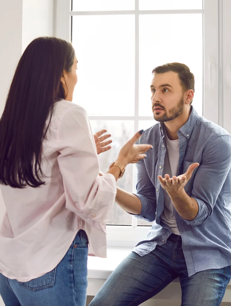 Pareja discutiendo frente a una ventana, mostrando tensión emocional y falta de comunicación como uno de los conflictos en pareja más comunes.