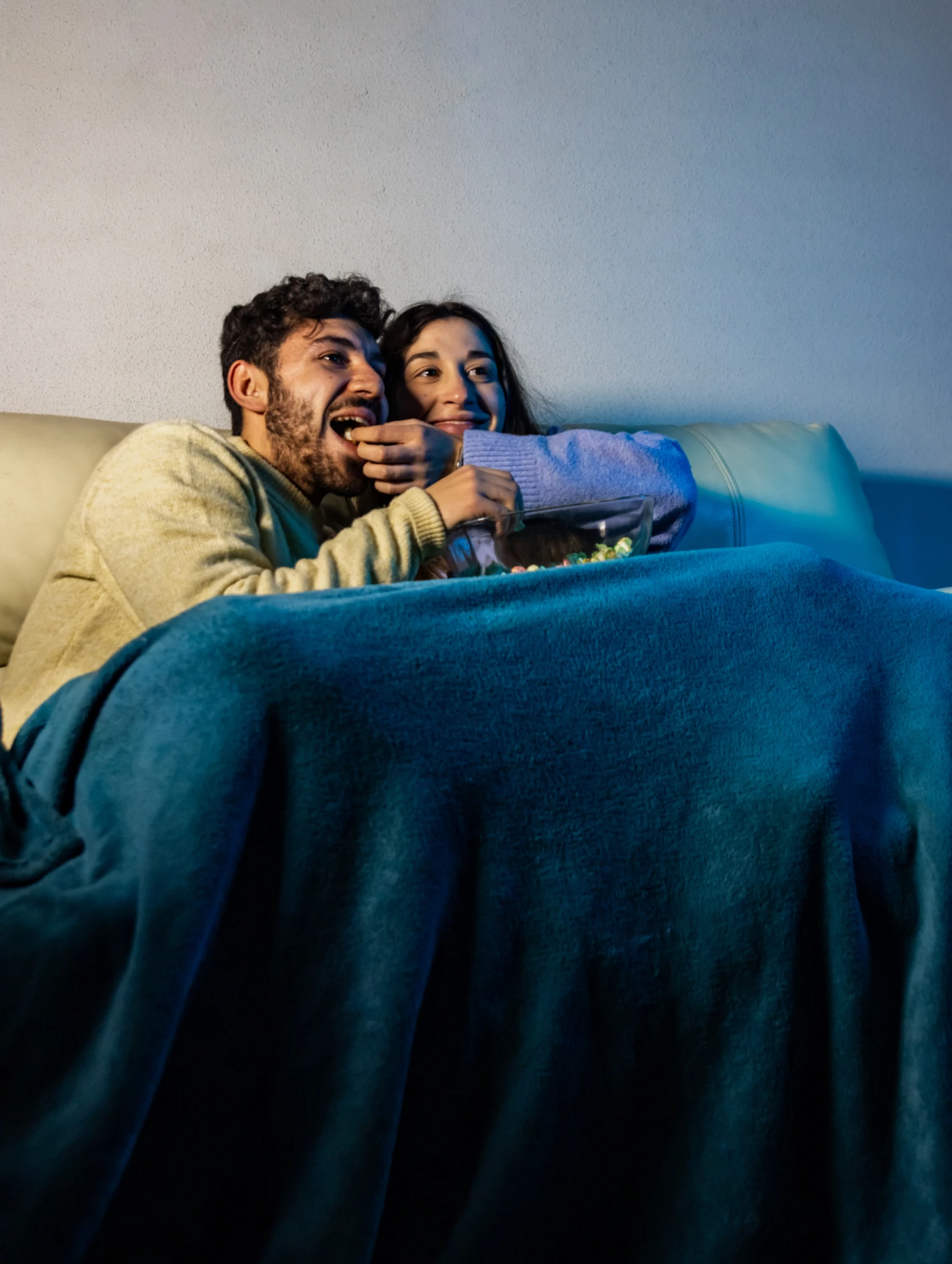 Pareja viendo una película en el sofá con manta y snacks, disfrutando de una noche acogedora de citas romanticas en casa con películas y momentos de conexión.