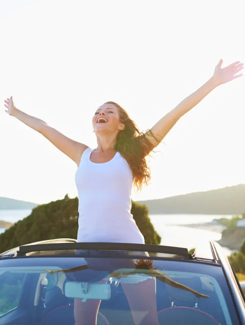 Mujer sonriente con los brazos abiertos en un auto descapotable junto al mar, satisfacción femenina y libertad al atardecer