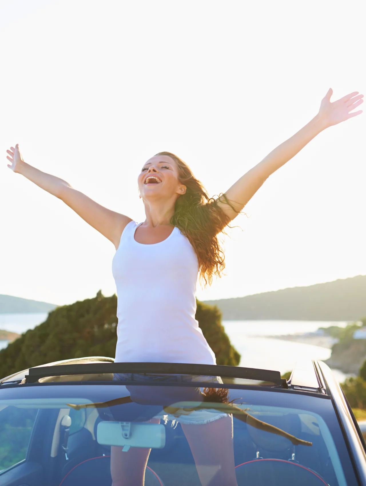 Mujer sonriente con los brazos abiertos en un auto descapotable junto al mar, satisfacción femenina y libertad al atardecer