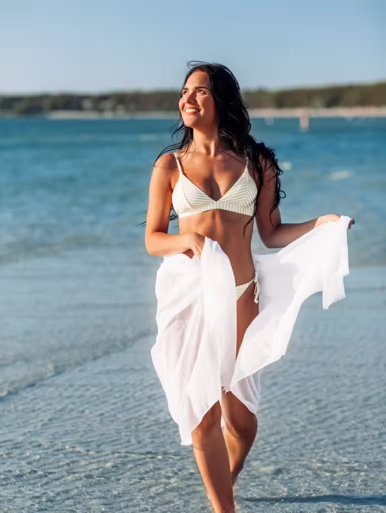 ¿Es normal tener granitos en la parte íntima La respuesta sin filtros que necesitas leer, mujer caminando en la playa con bikini claro y pareo blanco, es normal tener granitos en la parte intima